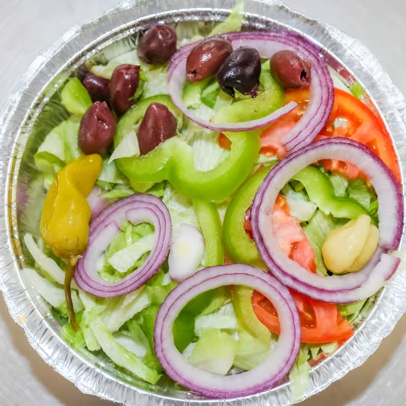 Fresh salad with lettuce, tomatoes, green peppers, red onions, olives, and a pepperoncini in a foil container.